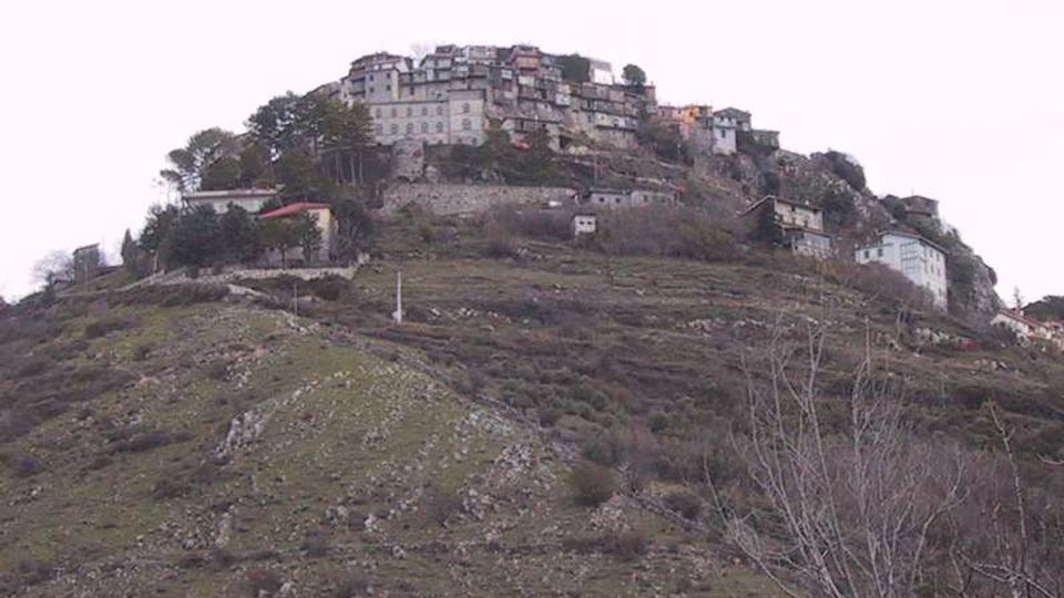 Borgo collinare con case in pietra addossate tra loro sulla sommità, circondato da mura e terrazzamenti. In primo piano pendio roccioso con vegetazione bassa e alberi sparsi.