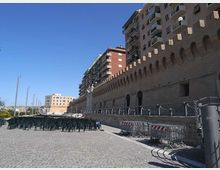 Ampia area pavimentata in pietra lungo una lunga muraglia in mattoni con arcate e merlature, affiancata da edifici residenziali alti con balconi. In primo piano si vedono file di sedie impilate, transenne metalliche e lampioni, sotto un cielo sereno.