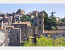 Veduta di un borgo storico con case in pietra e tetti in coppi, addossate su un pendio. Al centro spicca una torre campanaria in pietra con finestre ad arco, circondata da alberi e vegetazione.