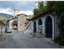 Strada di un borgo montano con edifici in pietra e una torre cilindrica sullo sfondo. A destra si vede un lungo edificio con portico ad archi e grate metalliche, mentre a sinistra ci sono case con balconi e la strada in lieve curva.