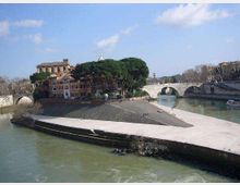 Veduta di un’isola fluviale alla confluenza di due rami del fiume, con viale alberato e edifici storici tra grandi pini. Sullo sfondo si vedono ponti in pietra ad archi che attraversano l’acqua lungo le banchine.