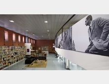 Interno di una biblioteca moderna con alte scaffalature di libri lungo pareti in mattoni e ampie finestre verticali. A destra corre un ballatoio curvo con grandi pannelli fotografici in bianco e nero, mentre al centro si trova un’area lettura con divani e tavolini su un tappeto.