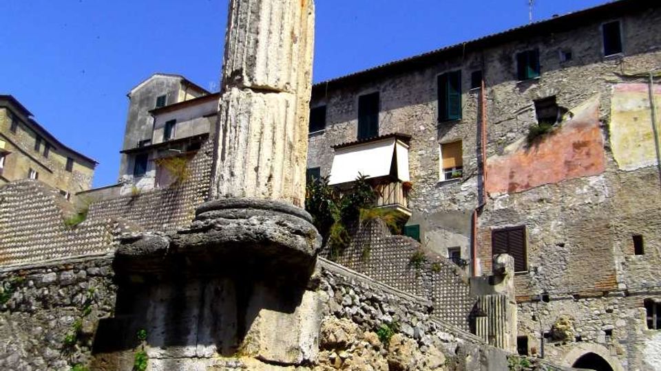 Colonna in pietra di epoca antica, parzialmente danneggiata e rinforzata con anelli metallici, su un basamento in muratura tra rovine e pietrame. Sullo sfondo si vedono edifici in pietra con finestre e persiane in un contesto urbano storico sotto cielo sereno.