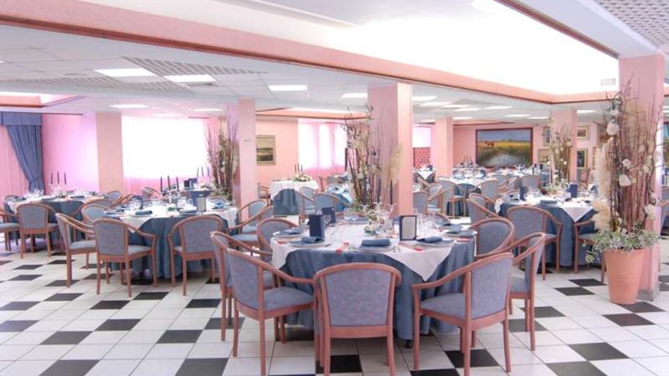 Ampia sala da pranzo interna con tavoli rotondi apparecchiati, sedie imbottite e decorazioni floreali alte. Pavimento a scacchiera bianco e nero, colonne rosa e soffitto con pannelli luminosi in un ambiente tipo ristorante o sala ricevimenti.