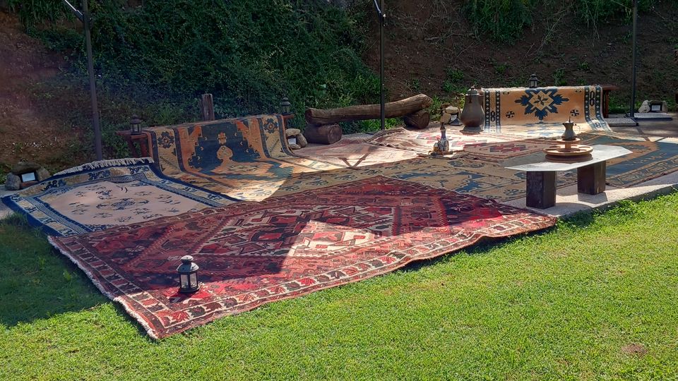 Area lounge all’aperto su prato con tappeti decorati e cuscini a terra, ombreggiata da grandi ombrelloni bianchi. Sullo sfondo ci sono una scarpata rocciosa e alberi, con piccoli tavolini bassi e lanterne.