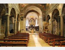 Interno di una chiesa in stile romanico con navata centrale e arcate su colonne in pietra, soffitti e pareti decorati con affreschi. Panche di legno disposte su entrambi i lati conducono verso l’altare in fondo, illuminato da una luce proveniente dall’abside.