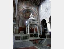 Interno di una chiesa in pietra con grande abside affrescata e arcate laterali; al centro un ciborio marmoreo a colonne sopra l’altare, con pavimento a motivi geometrici e copertura a capriate lignee.