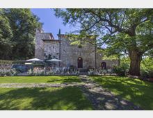 Edificio storico in pietra con facciata semplice, piccolo campanile e ingresso ad arco, affacciato su un giardino curato con prato, aiuole fiorite e tavoli all’aperto con ombrelloni. Grandi alberi e vegetazione fitta circondano l’area, con un sentiero che attraversa il prato.