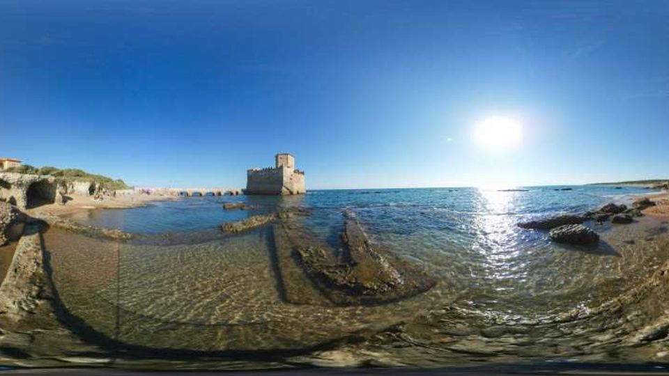 Panorama di una costa rocciosa con acqua bassa e trasparente; al centro si vede una piccola torre in pietra su uno scoglio vicino alla riva. Sulla sinistra ci sono scogli e vegetazione costiera, mentre a destra si estende il mare aperto con il sole basso che si riflette sull’acqua.