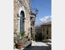 Vicolo in un borgo in pietra con scala in discesa, edifici in muratura e tetti in coppi. Sulla sinistra si vede una facciata con porta ad arco, vasi di piante e una lanterna a muro, con cavi sospesi e cielo azzurro sullo sfondo.