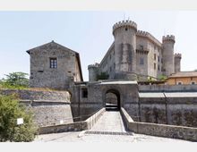 Castello in pietra con torri cilindriche merlate e mura fortificate, visto dall’ingresso con arco e ponte pedonale in ciottoli che conduce al portale. Sullo sfondo cielo sereno e vegetazione bassa ai lati delle fortificazioni.