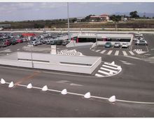 Ampio parcheggio asfaltato con numerose auto, corsie di ingresso e uscita segnalate da strisce pedonali e frecce direzionali. Al centro si vede una rampa di accesso a un parcheggio coperto con barriere e segnaletica, sullo sfondo campagna e colline.