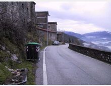 Strada asfaltata di montagna che curva lungo un muretto in pietra, con case in pietra addossate al pendio sul lato sinistro. Sullo sfondo si vedono vallate nebbiose e cime montuose, con un cassonetto verde in primo piano vicino al margine della strada.