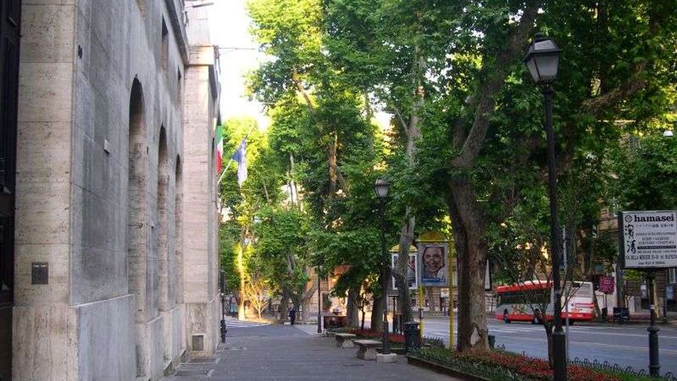 Marciapiede ampio lungo un edificio in pietra con grandi arcate, affacciato su un viale alberato con lampioni. Ai lati si vedono aiuole, panchine e la strada con veicoli sullo sfondo.
