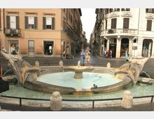 Fontana in pietra con vasca circolare e quattro sculture a forma di delfino che versano acqua, situata al centro di un incrocio tra strade lastricate. Sullo sfondo si vedono palazzi storici a più piani con persiane e negozi al piano terra lungo una via urbana stretta.