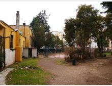 Cortile urbano con area sterrata e una rete metallica che delimita un piccolo campo sportivo; a sinistra si vede un edificio basso giallo con camino. Alberi e cespugli circondano lo spazio, con palazzi residenziali sullo sfondo.