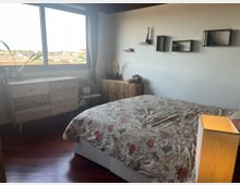 Camera da letto luminosa con grande finestra e tende grigie affacciata sui tetti della città, pavimento in legno scuro e letto con copriletto floreale. Sulla parete bianca ci sono mensole e cubi a muro, accanto a comò e credenza in legno decorato con piccoli oggetti e una lampada da tavolo.