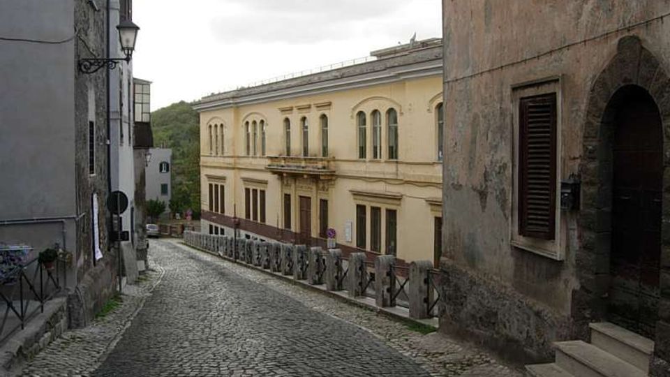 Strada in discesa lastricata in pietra tra edifici storici, con un grande palazzo chiaro a due piani con finestre ad arco sullo sfondo. Ai lati si vedono facciate intonacate, un lampione a muro e una ringhiera lungo il bordo della strada sotto un cielo nuvoloso.