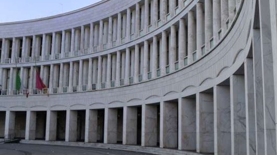Edificio monumentale a pianta semicircolare con facciata in pietra chiara, colonnato al piano terra e lunghe file di finestre verticali ai piani superiori. Davanti si apre una grande area pavimentata curva, con marciapiede e bandiere esposte sotto il portico.