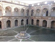 Cortile interno di un edificio storico in pietra, circondato da un porticato su due livelli con archi e colonne. Al centro si trova un pozzo con struttura metallica, su pavimentazione in ciottoli disposta a raggiera.