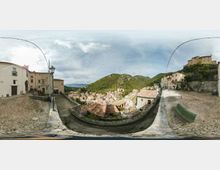 Veduta panoramica da una terrazza belvedere in pietra con pavimentazione a ciottoli, lampione e panchina, affacciata su un borgo di case in pietra con tetti in coppi. Sullo sfondo si vedono colline verdi e un castello o fortificazione sulla sommità, sotto un cielo nuvoloso.