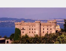Grande castello in pietra con mura merlate e torri angolari, circondato da vegetazione in primo piano. Sullo sfondo si vede un lago e colline lontane sotto un cielo limpido.