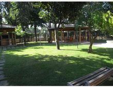 Giardino con ampio prato e alberi ombreggianti, con una panchina in primo piano e lampioni lungo il perimetro recintato. Sullo sfondo si vedono due strutture coperte in legno e tegole, una con area barbecue e l’altra con tavoli e sedie.