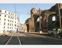 Strada urbana con binari del tram e cavi aerei, affiancata da un grande muro in mattoni con arcate in rovina. Sullo sfondo si vede un edificio bianco a più piani e l’area stradale con marciapiedi e cassonetti.