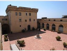 Cortile interno pavimentato in mattoni rossi, con un pozzo in pietra al centro e alcune fioriere. Sullo sfondo si vedono edifici color ocra con finestre rettangolari e un porticato ad archi lungo il lato destro.