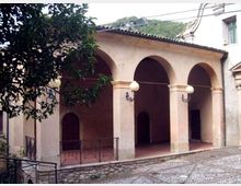 Portico di un edificio in muratura intonacata con tre grandi arcate e colonne squadrate, pavimento in cotto e lampioni a sfera fissati alle pareti. In primo piano si vede una piccola area lastricata con ringhiera metallica e un albero, con colline verdi sullo sfondo.
