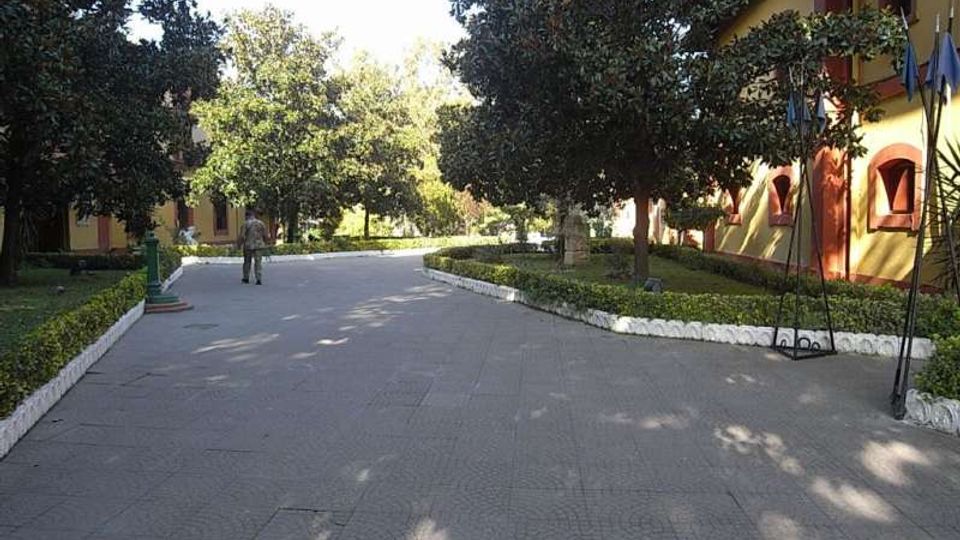 Viale pedonale pavimentato in un giardino, affiancato da siepi basse e grandi alberi che creano zone d’ombra. Sulla destra si vede un edificio con facciata giallo-ocra e finestre ad arco, con aiuole curate lungo il bordo.