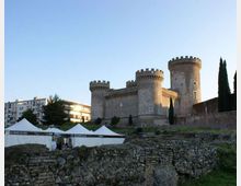 Castello in pietra con torri cilindriche merlate su un lieve rialzo, affiancato da cipressi e mura. In primo piano si vedono rovine in muratura e alcune tensostrutture bianche, con palazzi moderni sullo sfondo.
