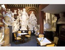 Interno di un locale simile a un caffè-museo con tavolini apparecchiati e sedie imbottite, circondato da numerose grandi statue e busti in gesso disposti lungo le pareti. Soffitto con travi a vista, colonne in legno e pareti ricoperte di sculture e pannelli informativi.