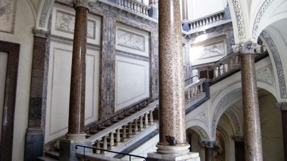 Atrio interno di un edificio storico con grandi colonne in pietra e archi a volta decorati. Una scalinata in marmo con balaustre conduce ai piani superiori illuminati da ampie finestre.