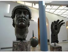 Sala museale luminosa con pareti bianche, lucernario sul soffitto e colonne, con grandi sculture scure su piedistalli in marmo, tra cui una testa monumentale e una mano. Sullo sfondo si vedono archi e altre opere esposte nello spazio.