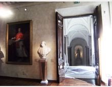 Interno di una sala museale con pareti chiare, un grande dipinto incorniciato e un busto su piedistallo. Una porta in legno aperta conduce a un corridoio con volte a crociera e pavimento a motivi geometrici, con una statua in fondo.