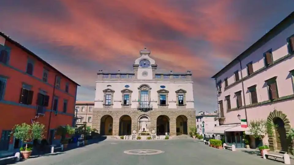 Piazza cittadina con al centro un edificio storico in stile classico, facciata bianca con portico ad archi al piano terra e balconi al piano superiore. La piazza è circondata da palazzi colorati e presenta una fontana centrale e vasi con piccoli alberi lungo i lati.