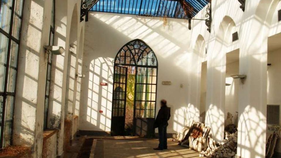 Interno di una galleria coperta con tetto in vetro e struttura metallica, pareti bianche con arcate e grandi finestre laterali. In fondo si vede un portale ad arco vetrato, con luce solare che proietta ombre geometriche sul pavimento.