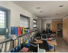 Interno di un ufficio allungato con scaffalature piene di libri e raccoglitori lungo il lato sinistro sotto finestre con veneziane. Sulla destra ci sono postazioni con scrivanie, computer e sedie, armadi alti in legno e illuminazione a faretti incassati nel soffitto.
