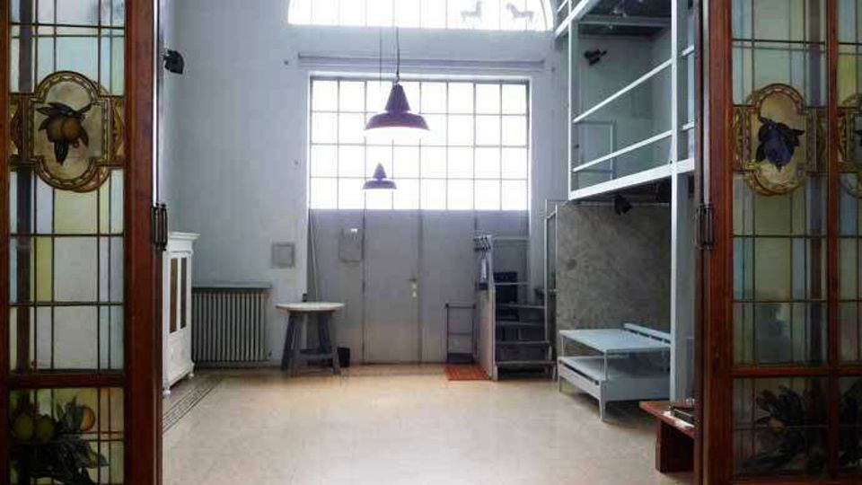 Interno di un ampio spazio tipo loft con grandi finestre ad arco e a griglia sopra una porta centrale, illuminato da due lampade a sospensione. Ai lati ci sono porte in legno con vetri decorati, una scala metallica che porta a un soppalco e arredi essenziali su pavimento chiaro.
