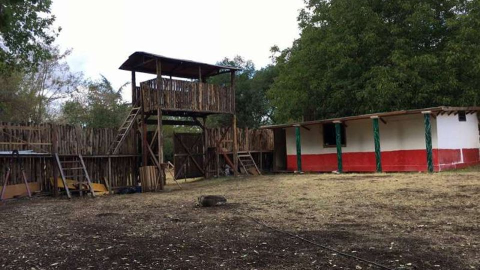 Cortile rurale recintato con una torre di osservazione in legno sopraelevata con scala e piattaforma coperta. A destra c’è un edificio basso bianco con fascia rossa e colonne verdi sotto una tettoia, circondato da alberi.