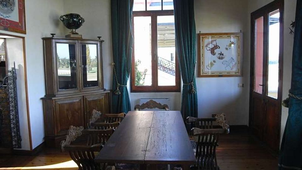 Sala da pranzo con lungo tavolo in legno e sedie intagliate, pavimento in legno e credenza vetrata. Una grande finestra con tende verde scuro illumina l’ambiente, con quadri alle pareti e porta finestrata sul lato destro.
