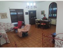 Soggiorno-sala da pranzo con pavimento in cotto, tavolo in legno con sedie e credenza scura contro la parete. Sono presenti un divano con copridivano decorato, poltrone, lampadario a soffitto e due grandi aperture con infissi in legno (porta-finestra e finestra ad arco).