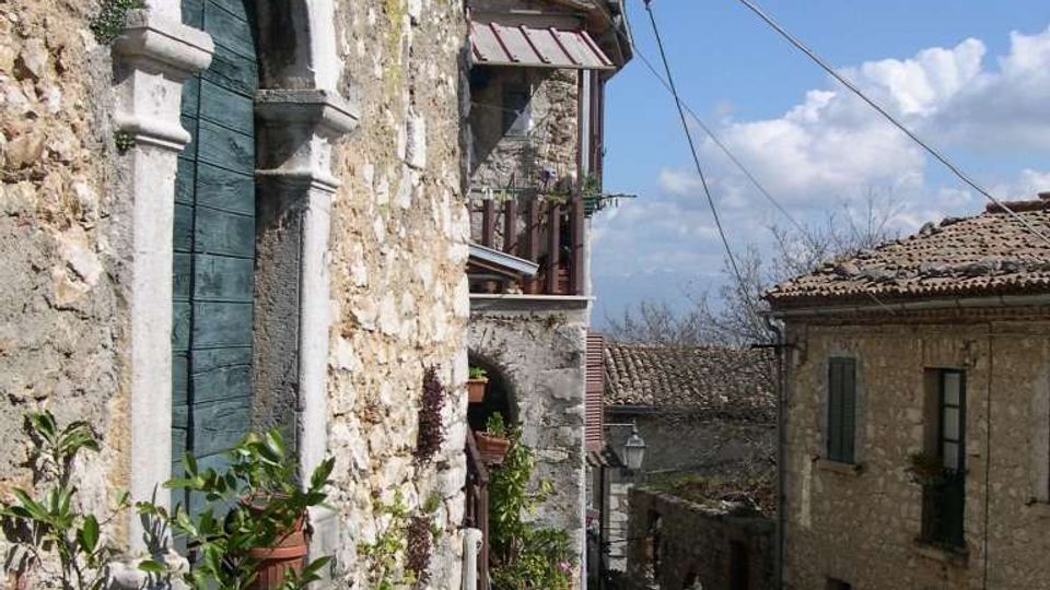 Vicolo in un borgo in pietra con scala in discesa, edifici in muratura e tetti in coppi. Sulla sinistra si vede una facciata con porta ad arco, vasi di piante e una lanterna a muro, con cavi sospesi e cielo azzurro sullo sfondo.