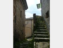 Vicolo stretto tra edifici in pietra con pavimentazione irregolare, illuminato da un lampione a parete. A destra una scalinata in pietra sale tra muretti con vegetazione spontanea, con ringhiera e piccola tettoia in alto.