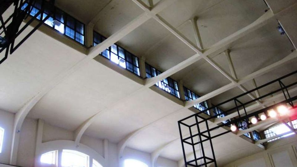 Vista verso l’alto dell’interno di un grande edificio industriale con soffitto alto a capriate e travi in cemento, finestre a nastro lungo la sommità e archi sulle pareti. Sono presenti strutture metalliche a griglia e un’illuminazione con faretti sospesi.