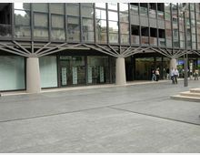 Facciata di un edificio moderno con ampie vetrate a griglia e colonne cilindriche che sorreggono una struttura a travi incrociate. Davanti si apre una grande piazza pavimentata in cemento, con ingresso coperto e vetrine al piano terra.