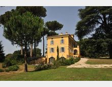 Villa di campagna a tre piani intonacata di giallo, con finestre a persiane e ingresso ad arco, circondata da un ampio giardino con pini alti. Un vialetto sterrato conduce all’edificio, con prato e alberi sullo sfondo.