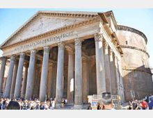 Facciata di un tempio romano con alto portico a colonne corinzie e frontone triangolare in pietra, con iscrizione latina. Sullo sfondo si vede la grande rotonda in muratura della struttura, in una piazza urbana.