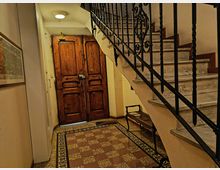 Ingresso interno di un edificio con grande portone in legno e pavimento a scacchiera con bordo decorato. A destra una scala in marmo con corrimano in ferro battuto e una piccola panca sotto le scale.